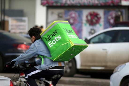 Person wearing a jean jacket riding a bike with a green "Uber Eats" bag on their back.