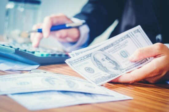 Person using a calculator with one hand and hundred dollar bills in the other hand.