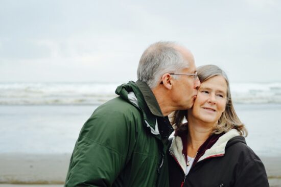 Older man kissing an older woman with the beach behind them.