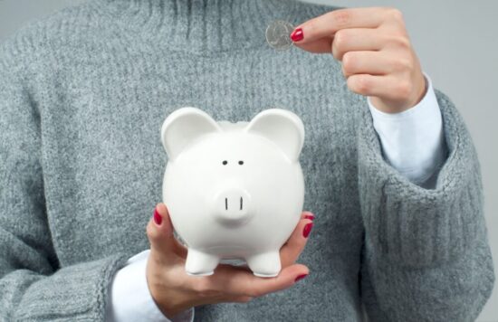 Person in a gray sweater holding a white piggy bank and a coin they're about to add to the bank.